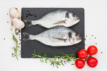 Fresh dorado fish on slate cutting board, rosemary, garlic and tomatoes on white table. Top view, copy space.