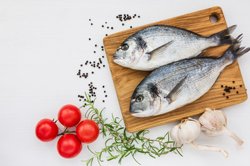 Fresh dorado fish on wooden cutting board with garlic, rosemary and tomatoes on white table. Top view, copy space. 