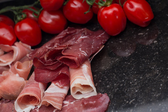 Jambon mix. Ham. Traditional Italian and Spanish salting, smoking, dry-cured dish - jamon Serrano and prosciutto crudo sliced with herbs and tomatoes on dark stone background. Copy space. Closeup. 
