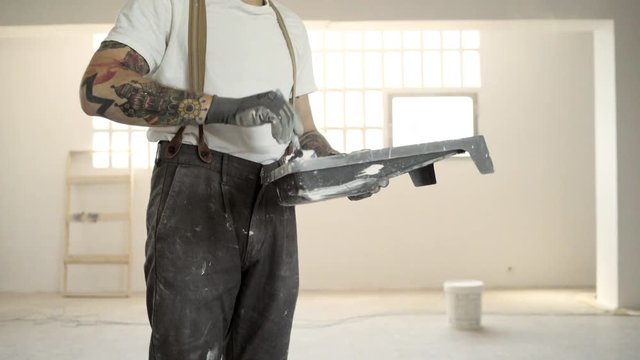 Closeup, unrecognizable tattooed man wered pants with suspenders is preparing to paint wall with brush and white paint Home diy renovation