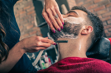 Female barber shaving a client's beard in a barber shop. Close-up