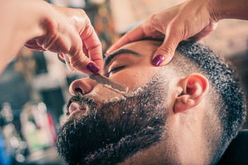 Female barber shaving a client's beard in a barber shop. Close-up
