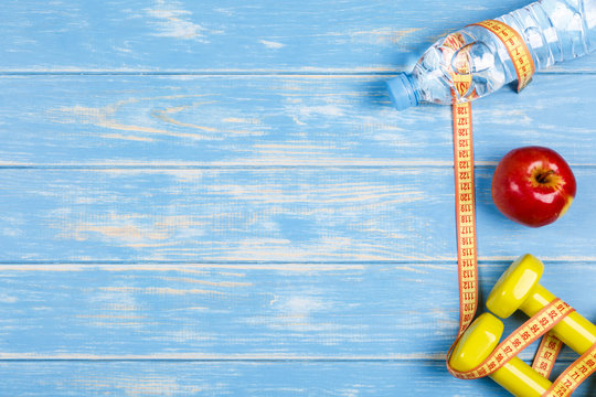 Bottle of water, apple, dumbbells and measure tape on blue wooden background.
