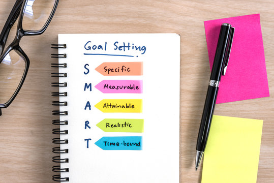 Hand writing definition for smart goal setting on notebook with pen eye glasses and colorful sticky note on desk