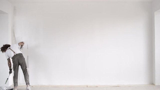 Funny curly man wears pants with suspenders quickly finishes to paint big wall from plasterboard with white paint and brush roller Home dyi renovation