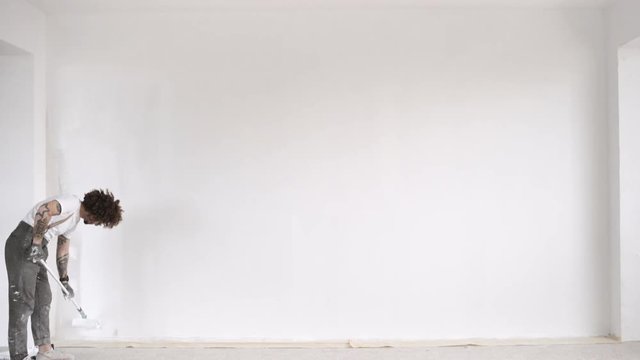 Funny curly man wears pants with suspenders finishes to paint part of big wall from plasterboard with white paint and brush roller Home dyi renovation Loopable video.