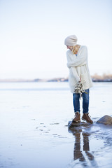 Closeup of beautiful young woman in warm clothes standing on a stone in water by the river shore. Girl enjoying winter sun and having fun outdoors. Lifestyle and winter concept.