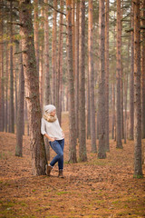 Fototapeta premium Portrait of beautiful woman standing at the old pine tree in the forest and enjoying sunlight. Girl walking in the park outdoors. Cold season. Lifestyle concept