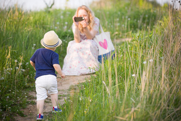 Mother taking photo of her adorable little toddler boy with smartphone. Family walking in the park. Small child with mom.