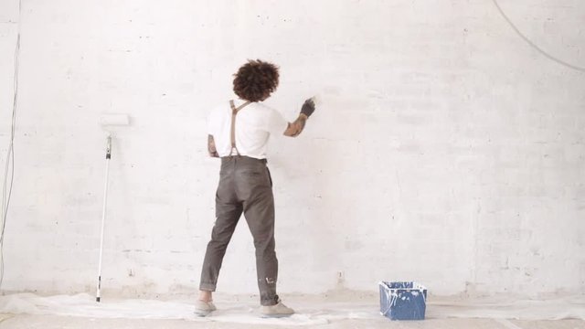 Funny curly man wears pants with suspenders carefully paints holes in brick wall with painting brush and white paint Home diy renovation