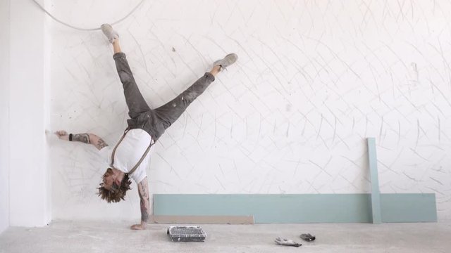 Funny curly man wears pants with suspenders makes sport exercises white painting wall, one hand headstand
