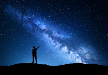 Fotobehang Milky Way. Silhouette of a standing man pointing finger in night starry sky on the mountain. Colorful night landscape with beautiful universe. Travel background with blue sky full of stars  © den-belitsky