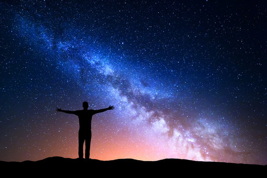 Night landscape with Milky Way. Silhouette of a standing young man with raised up arms on the mountain. Beautiful Universe. Travel background with blue night starry sky