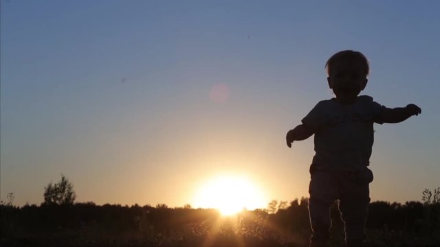 A little 1 year old baby boy is walking on sunset in nature. Child make his first steps outdoor. Kids Silhouette in the field with sundown