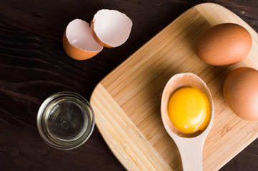 Chicken eggs on a wooden cutting board and egg yolk in a wooden spoon on a dark wooden table.