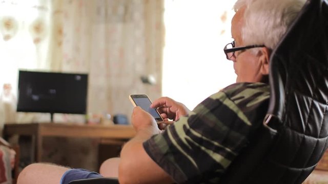 Senior man sitting with a phone in a leather chair near the house TV. Man typing sms and watching photo