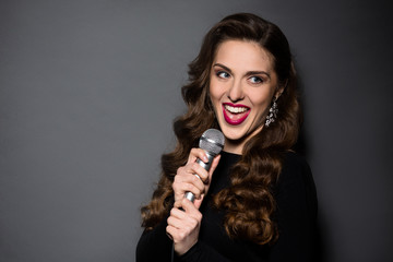 Beautiful lady with red lips singing in studio. Professional musician in black dress smiling and posing for photographer over grey background.