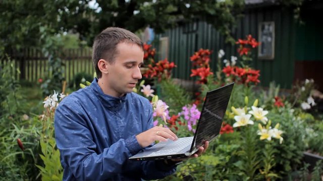 Young man working at a laptop near beds of flowers. White, red and purple flowers in the background