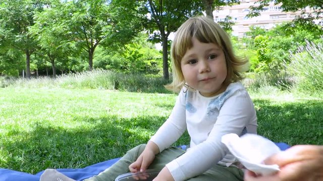 little blonde cute kid sitting in towel on grass at urban park eating with hand chicken and mushroom pieces in plastic tupperware
