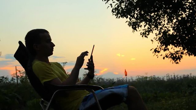 Silhouette of male in chair playing in tablet. Against the background of an orange sky after sunset