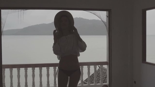 Pretty naturally looking woman on summer day in boho style fashion wearing white shirt, bikini and hat dancing in a abandoned building over beautiful sea landscape background.
