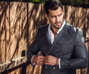 Elegant handsome man in classical grey suit poses near wooden fence. - 115956281