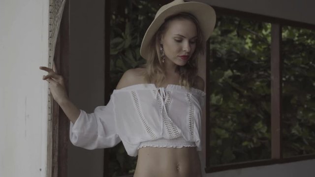 Pretty natural woman on summer day in boho style fashion wearing white shirt, bikini and hat walking in a abandoned building and looking through the window over beautiful green leaves background.