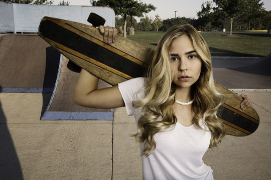Beautiful blond girl wearing white shirt and jean shorts standing with skateboard in a skate park.