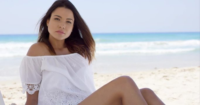 Gorgeous young woman relaxing on the beach sitting under the shade of an umbrella in a trendy white summer top looking at the camera