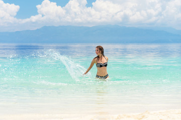 Fototapeta na wymiar Young european woman in bikini with good mood splashing and dancing in beautiful tropical calm sea under cloudy soft sky
