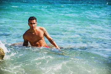 Attractive young shirtless athletic man sitting in water by sea or ocean shore, wearing shorts, looking at camera