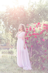 Fototapeta premium beautiful sexy girl in a light dress with delicate make-up and hair in a flower garden with roses in luchas sunlight at sunset. Styled photo fane art