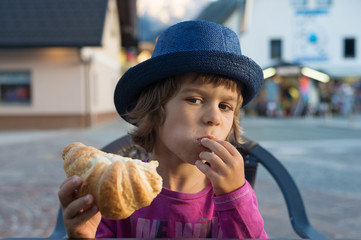 Cute little boy with blue hat sitting at the table stuffing croissant into his mouth.