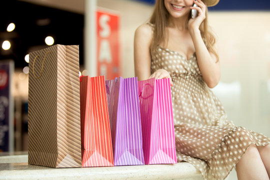 Close-up of beautiful smiling young shopper woman sitting with shopping paper bags talking on phone, making call. Focus on bags. Fashion, sale, shopping concept. Sale sign on background