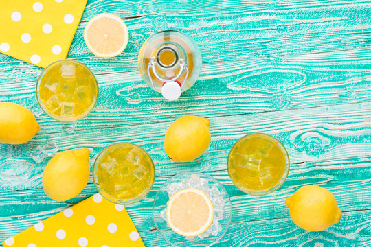 lemonade or limoncello in glasses with ice cubes, sherbet glass with ice cubes, bottle with drink, lemon fruits on turquoise colored wooden table decorated with yellow napkins at polka dots top view