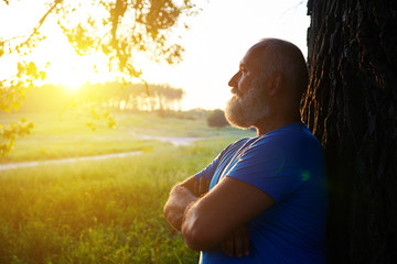 Naklejka na ściany i meble Side view of handsome aged man near the tree watching sunset