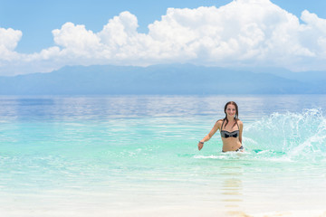 Young european woman in bikini with good mood splashing and dancing in beautiful tropical calm sea under cloudy soft sky