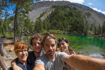 Four young people taking selfie in idyllic landscape with green lake, conifer woodland and...
