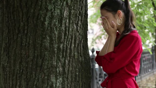 Young girl in a red dress crying near a tree in a city park