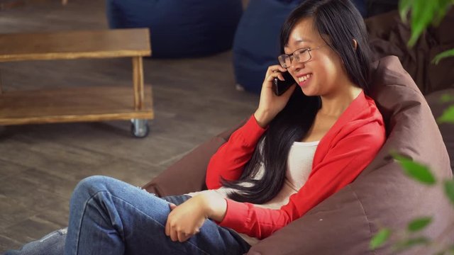 Asian woman talking on the mobile phone indoors. Young girl talks on the smartphone and smiling. Happy businesswoman using app fom calling. Casual interior. Brunette with long hair and eyeglasses