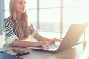 Young beautiful female copywriter typing texts and blogs in spacious light office, her workplace, using pc keyboard. Busy woman working.