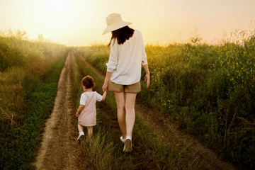 Amazing, charming, adorable view of a pretty mother in a hat and little beautiful daughter walking on a path of a field. Sunset or sunrise. Motherhood and childhood