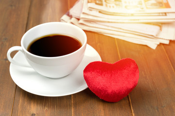 Coffee cup and heart with newspaper in background on wooden table. Sunlight filter effect photo.