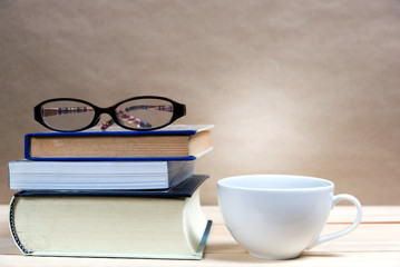 Stack of books with coffee cup and glasses on wooden table.