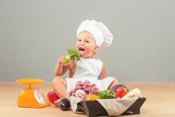 Little girl dressed in white chef hat and apron, eats only vegetables. The child weighed on the scales Products: eggplant, onion, garlic, pumpkin, red and green peppers, cabbage, tomatoes