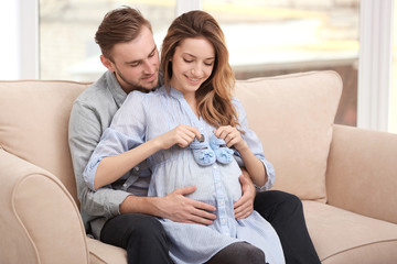 Happy pregnant woman holding baby shoes  and her husband on a sofa at home