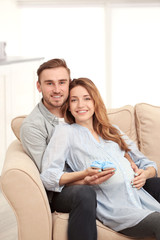 Happy pregnant woman holding baby shoes and her husband on a sofa at home