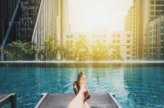 Relaxing In Holidays, Asian Guy Feet On Sun Bathing Bed At Roof Top Swimming Pool In The City