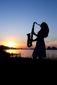 silhouette of a girl in a dress at dawn near the river in the village playing saxophone on a background of blue sky