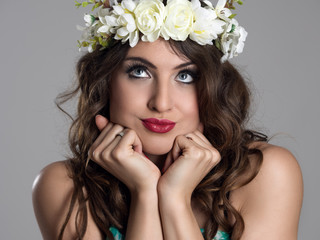 Cute young pensive beauty model wearing floral wreath with head in hands looking up over gray studio background